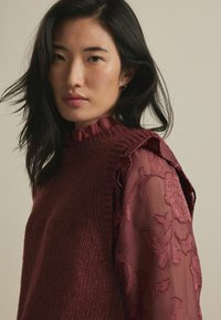 Pull en tricot bourgogne avec un col à volants et des manches transparentes, brodées de fleurs ; tissu texturé et détails de conception en couches.