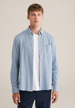 Hellblaues Button-Down-Hemd aus texturiertem Stoff, mit einer Brusttasche und einem lässigen Kragen, das über einem weißen T-Shirt getragen wird.