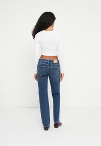 Blue denim jeans with a straight-leg fit, featuring two back pockets and a beige brand patch at the waistband. Paired with a white long-sleeve top.