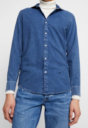 Mand iført blåt ribbet skjorte med knapper over en hvid rullekrave og lyseblå jeans, stående mod en ensfarvet baggrund.