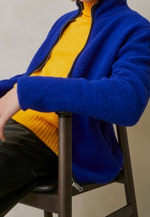 Giacca in pile blu con colletto alto e zip, indossata sopra un maglione giallo brillante a costine. I pantaloni in pelle nera creano un contrasto.