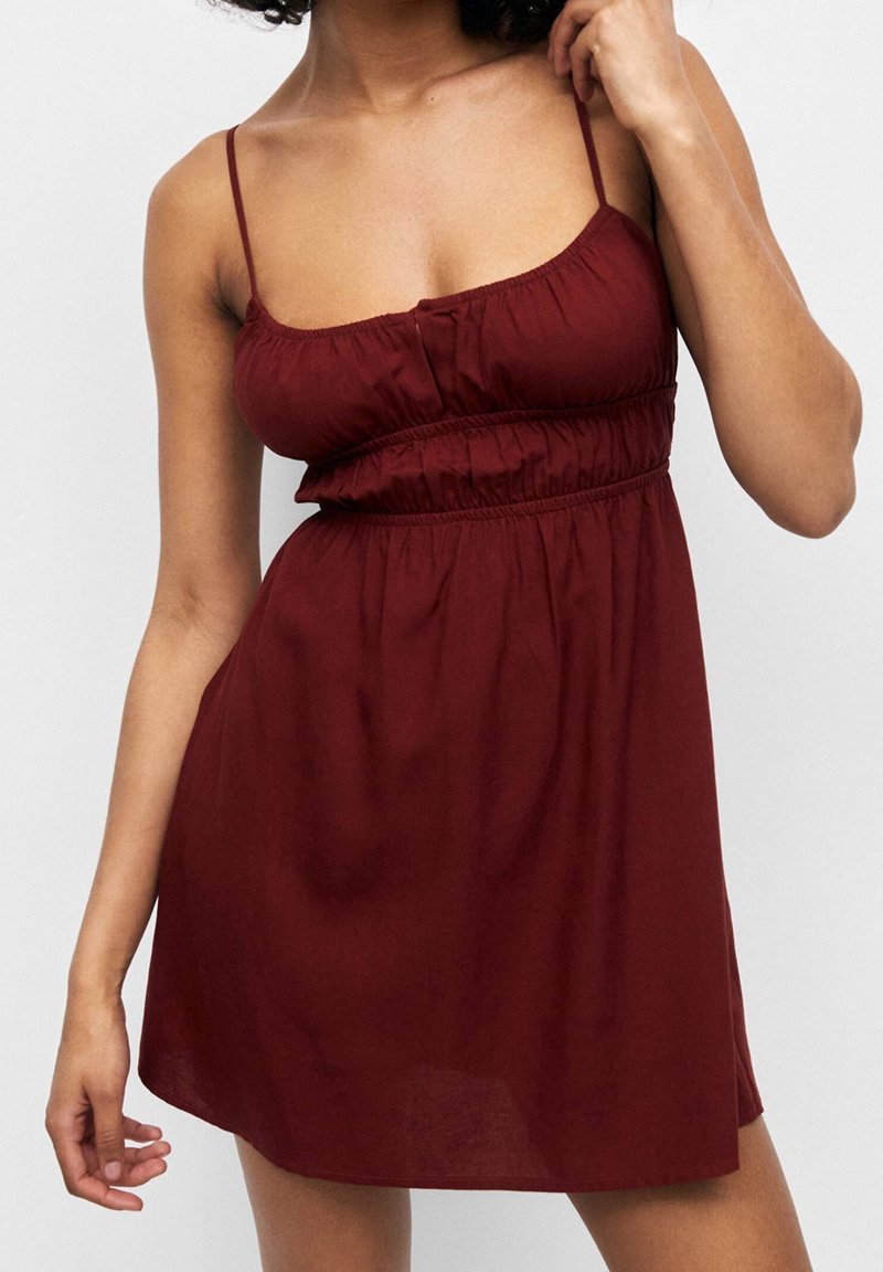 Vínové šaty bez rukávů s přiléhavým živůtkem a řaseným pasem, vyrobené z lehké látky, s tenkými ramínky a splývavou sukní.