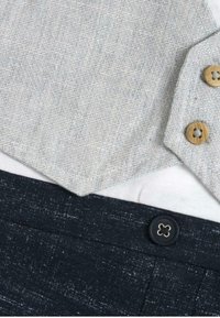 Chemise texturée gris clair avec des boutons en bois, superposée sur une chemise blanche. Pantalon bleu marine foncé avec un bouton décoratif et un motif subtil.