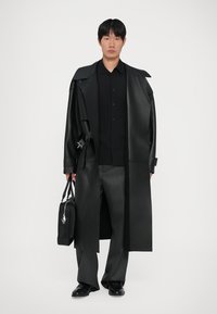 Long black leather trench coat with oversized collar, paired with a black button-up shirt, gray trousers, and holding a black bag.