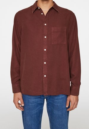 Mand iført en brun langærmet skjorte med brystlomme og blå jeans, stående med hænder afslappet langs siden.