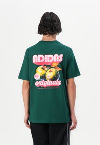 Zaļš kokvilnas T-krekls ar lielu grafiku, kurā attēloti divi āboli un uzraksts "ADIDAS originals" rozā un baltā krāsā. Īsas piedurknes, brīvs griezums.