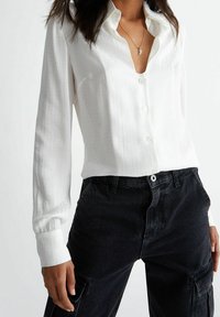Camicia bianca a maniche lunghe con scollatura a V profonda, caratterizzata da un sottile motivo testurizzato, abbinata a pantaloni cargo neri a vita alta.