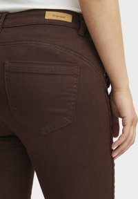 Jeans en denim marron foncé avec une texture lisse, présentant une poche arrière et une étiquette beige portant le nom "fransa". Design slim fit visible.
