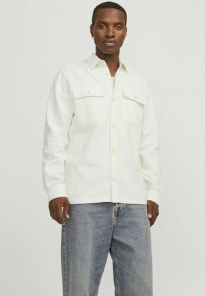 White button-up shirt with two chest pockets, long sleeves, and a relaxed fit, paired with light blue jeans and a plain undershirt.