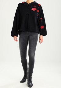 Person som bär en svart huvtröja med röda läppar och stjärnpatchar, kombinerad med mörkgrå skinny jeans och svarta ankelboots.