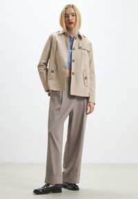 Giacca corta beige con due tasche frontali, bottoni marroni e colletto. Abbinata a pantaloni larghi grigi chiari e mocassini neri.