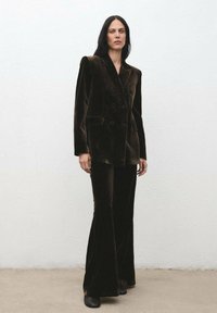 Blazer de terciopelo verde oscuro, de doble botonadura y con solapas de muesca, combinado con pantalones acampanados a juego. Textura suave y corte ajustado, botas de tobillo negras.