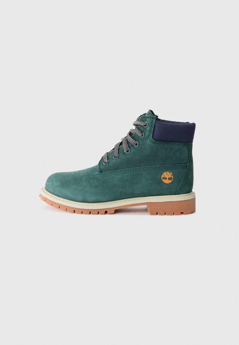 Bottine en suède vert avec lacets noirs et blancs, col rembourré bleu marine, semelle en caoutchouc beige et logo orange en forme d'arbre sur le côté.