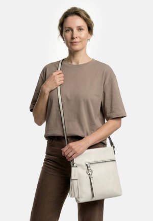 Woman wearing taupe shirt and brown pants, holding white crossbody bag with tassel and zipper details, standing against white background.