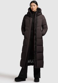 Tumepruun pikk puffertak someday koos kapuutsiga, esise eestluku ja suurte tüüblitega. Kannab musta riietuse peal.