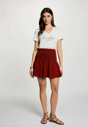 Donna con capelli lunghi e scuri che indossa una maglietta bianca con la scritta "Paradise Island", gonna rossa, gioielli dorati e sandali marroni borchiati, in piedi davanti a uno sfondo semplice.