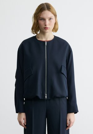 Νεαρό άτομο με ξανθά μαλλιά έως τους ώμους, φοράει απλό μαύρο μπουφάν με φερμουάρ και ταιριαστό μαύρο παντελόνι, στέκεται μπροστά σε λευκό φόντο.