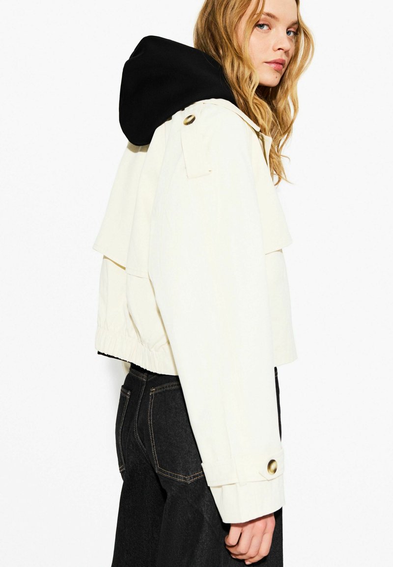 Veste crème avec une capuche noire, design court, taille élastique et accents de boutons dorés. Texture lisse avec un devant superposé.
