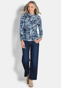 Langärmlige blaue Bluse mit Blumenmuster und Stehkragen, kombiniert mit dunkelblauen weit geschnittenen Jeans und beigen Stiefeletten. Strukturiertes Blumenmuster.