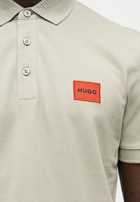 Φωτεινό μπεζ πόλο με τρία λευκά κουμπιά και μια κόκκινη ορθογώνια ραφή με την επιγραφή "HUGO" στο αριστερό στήθος.