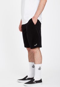 Pantaloni corti in corduroy neri con tasche laterali, una vestibilità comoda e ricamo del logo bianco. Indossati con scarpe slip-on nere e calzini bianchi a motivi.