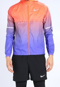 Chaqueta cortavientos Nike con degradado de naranja a púrpura, tejido ligero, cierre de cremallera y detalles reflectantes. Combinada con pantalones cortos negros.