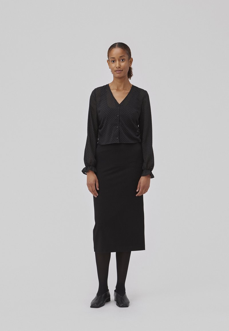 Blouse noire à boutonnières avec un motif texturé, assortie à une jupe crayon noire jusqu'aux genoux. Des collants noirs et des chaussures à bout pointu complètent le look.