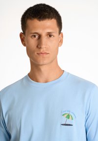 Vaaleansininen pitkähihainen paita, joka on valmistettu pehmeästä kankaasta ja jossa on vasemmassa rinnassa brodeerattu merenrantateema, jossa on auringonvarjo.