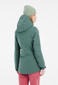 Grüne wasserdichte Jacke mit Kapuze, elastischen Bündchen und einem schlichten Design. Kombiniert mit rosa Hosen mit Reißverschlusstaschen.