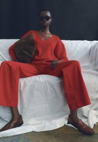 Maglione rosso a maglia con scollo a V, pantaloni rossi larghi abbinati, borsa a tracolla in suede marrone e mocassini marroni, su uno sfondo bianco drappeggiato.