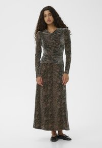 Frau mit langen, lockigen Haaren, die ein langes, gerafftes, Tierdruckkleid und schwarze flache Schuhe trägt, steht vor einem schlichten Hintergrund.