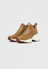 Rjave superge z vpikanjem iz pletenega materiala in teksturirano zunanjo površino. Imajo zadrgo, masivni beli podplat in tkaninske poudarke.