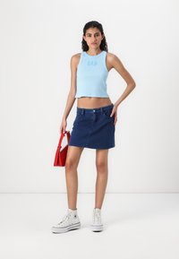 Lyseblå, kort tanktop med "GAP"-logo, kombinert med et mørk denimbbrevkjole. Modellen holder en rød håndveske. Hvite høye sneakers.