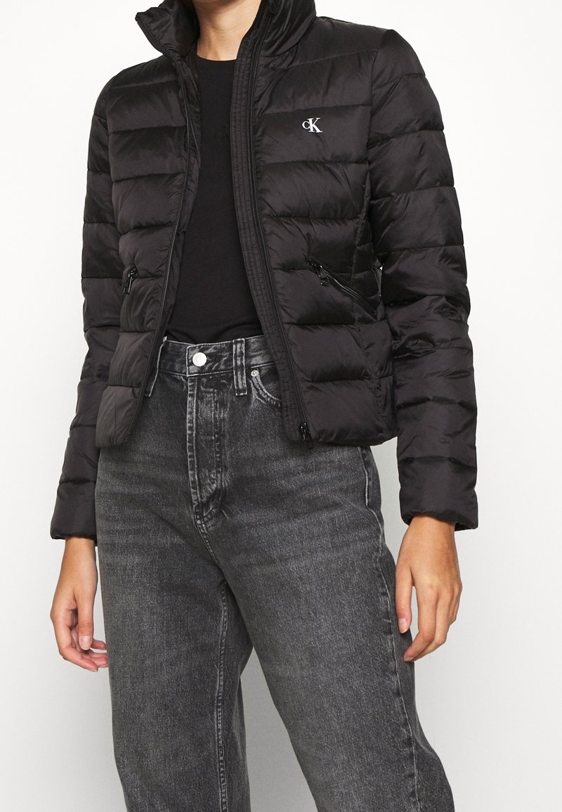 Personne portant une veste matelassée noire Calvin Klein par-dessus une chemise noire, associée à un jean noir délavé taille haute.