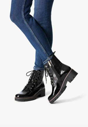 Jambes portant un jean bleu ajusté et des bottines noires brillantes à lacets avec fermetures éclair, semelles épaisses et talons bas sur fond blanc.