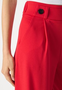 Czerwone dopasowane spodnie o gładkiej fakturze, z plisowanym przodem i czarnym zapięciem na guzik. Widoczna ręka trzymająca w talii.