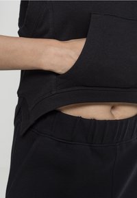 Zwarte cropped sweatshirt met een voorzak en geribbelde onderrand, gecombineerd met zwarte high-waisted joggingbroeken. De gladde stoftextuur is zichtbaar.