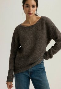 Brązowy sweter dzianinowy z szerokim dekoltem, luźnym krojem i długimi rękawami, zestawiony z niebieskimi dżinsami. Teksturowana tkanina z widocznymi detalami przeszycia.