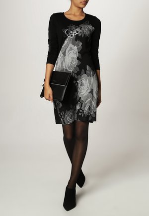 Femme portant une robe longue à manches noires avec un motif floral argenté, des collants noirs, des chaussures noires à bouts pointus, tenant une pochette noire.