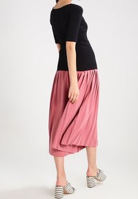 Schwarzes figurbetontes Oberteil mit Dreiviertelärmeln, kombiniert mit einem plissierten rosa Midikleid. Das Modell trägt gestreifte Blockabsatzsandalen.