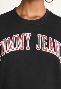 Mustapaita, jossa on suuri punainen "TOMMY JEANS" -teksti, ribattu pyöreä kaula-aukko ja pehmeä kangas.