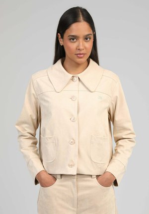 Femme portant une veste beige boutonnée avec un large col et un pantalon assorti, debout les mains dans les poches devant un fond uni.