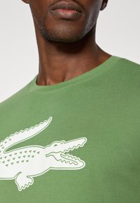 Zaļš kokvilnas T-krekls ar lielu baltu krokodila logotipu, apkakles dizains un gluda tekstūra. Tuva skata skats uz apkakles un krūšu daļu.