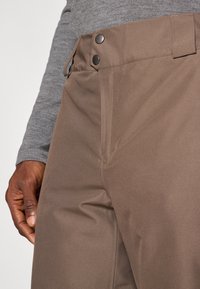 Brown fabric trousers featuring a smooth texture, two metal snap buttons at the waist, and clean lines tailored for a fitted look.
