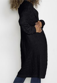 Czarny dzianinowy kardigan o otwartej teksturze, z długimi rękawami i prążkowanymi mankietami, charakteryzujący się luźnym, wydłużonym fasonem.