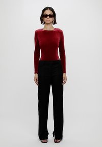 Roter Samt-Bodysuit mit langen Ärmeln, kombiniert mit schwarzen hoch taillierten Hosen. Das Model trägt dunkle Sonnenbrillen und große Creolen. Einfache Hintergrund.