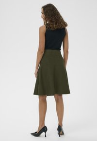 Gonna A-line verde oliva con vita alta aderente, realizzata in tessuto testurizzato. Indossata con un top senza maniche nero e tacchi neri.