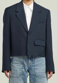 Blazer navy ajustado hecho de tela suave, con cuello con muesca, un solo botón y bolsillo de parche frontal, usado sobre una camisa blanca.
