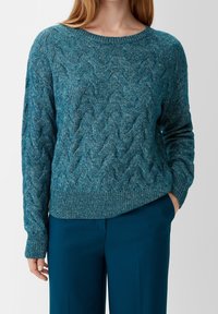 Frau, die einen türkisfarbenen Kabelstrickpullover mit gerippten Bündchen und Saum trägt, kombiniert mit dunkel-türkisen Hosen, die Hand in der Tasche, vor einem einfarbigen Hintergrund.