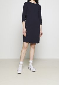 Marinblå klänning i bomull med trekvartslånga ärmar, rund halsringning med kontrasterande kant, sidofickor och en liten logotypdetalj nära fållen.